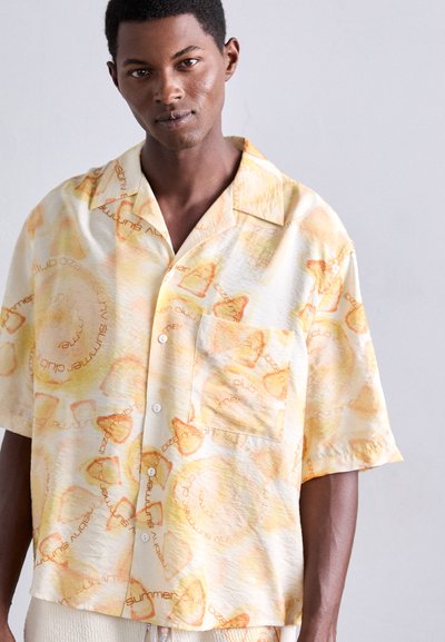 Un jeune homme portant une chemise à manches courtes avec des motifs abstraits jaunes et oranges et du texte, se tenant devant un fond clair.