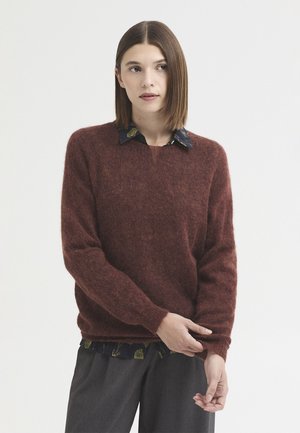 Jeune femme aux cheveux bruns et raides porte un pull marron doux par-dessus une chemise sombre à motifs et un pantalon gris foncé, ajustant sa manche.