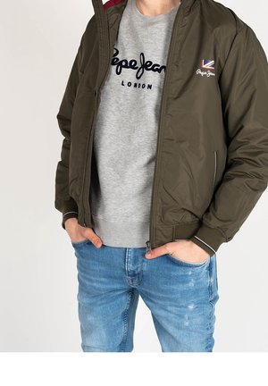 Personne portant une veste bomber vert olive dézippée sur un sweat-shirt gris Pepe Jeans London et un jean bleu avec les mains dans les poches.