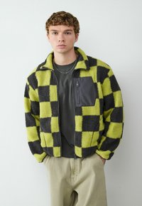 Pluche geruit jack in groen en zwart, met een ritszak op de borst en hoge kraag. Draag het over een donker shirt en lichte broek.