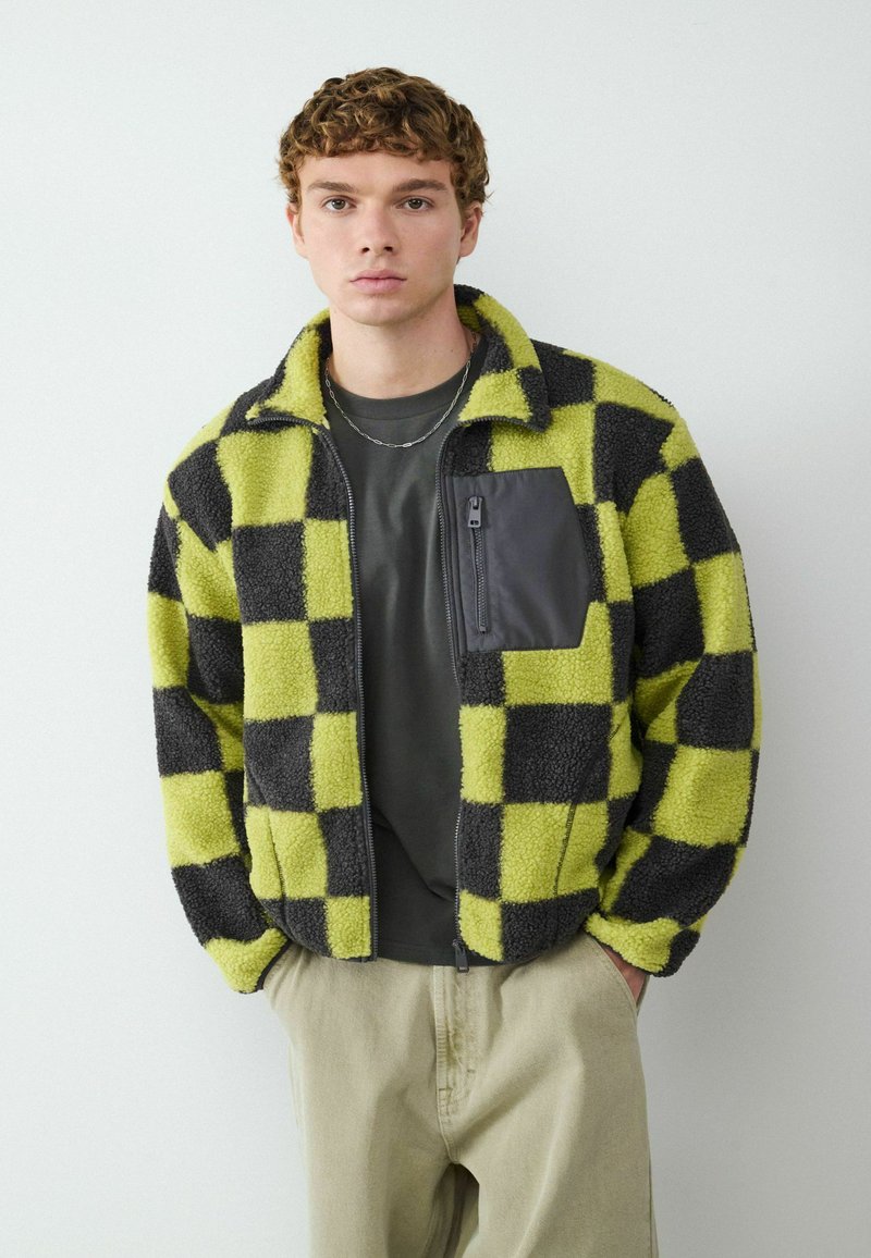 Pluche geruit jack in groen en zwart, met een ritszak op de borst en hoge kraag. Draag het over een donker shirt en lichte broek.