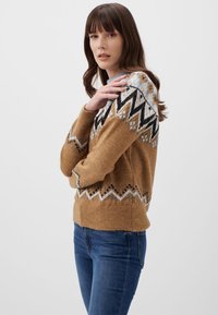 Maglione lavorato a maglia marrone con motivi geometrici in bianco e nero, scollo rotondo e maniche lunghe, abbinato a jeans blu.