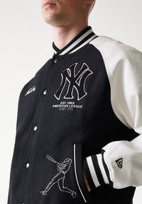 Μαύρο πουλόβερ varsity από μαλλί με λευκά δερμάτινα μανίκια, διαθέτει κεντημένο λογότυπο NY, γραφική απεικόνιση μπέιζμπολ και ριγωτές ριμπ μανσέτες.