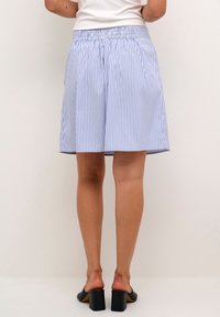 Žena oblečená v sukni s modro-bílými svislými pruhy, bílém topu a černých lodičkách na podpatku, stojící proti jednoduchému světlému pozadí.