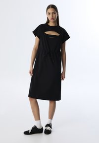 Schwarzes Baumwollkleid mit kurzen Ärmeln, das über einen Ausschnitt mit Cut-Out und eine verstellbare Kordelzugtaille verfügt. Sanfte Textur, knielanges Design.