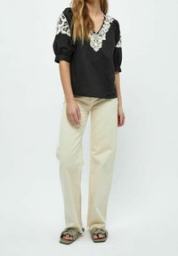 Chemisier noir avec des détails en dentelle sur les épaules et le décolleté, associé à un jean beige à jambes larges et des sandales vertes à glisser.