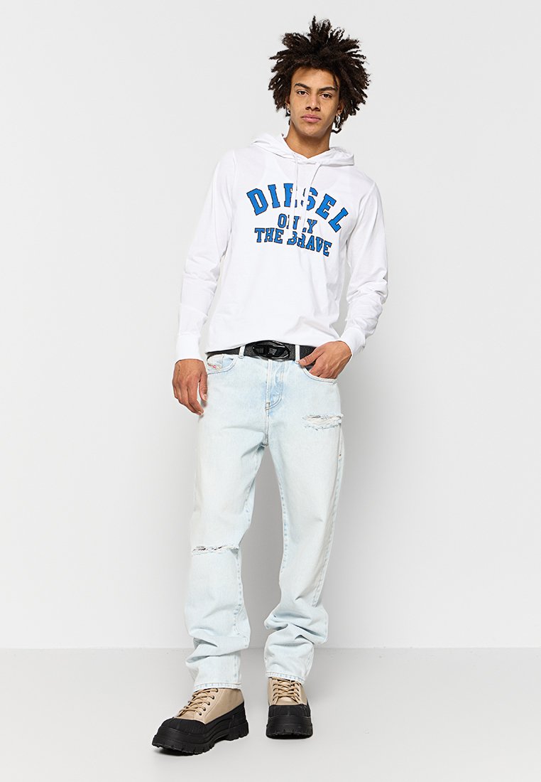 Weißer Kapuzenpullover mit blauer Druckschrift, kombiniert mit hellblauen, abgetragenen Jeans und beige-schwarzen, klobigen Stiefeln.