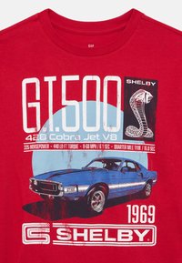 Κόκκινο βαμβακερό T-shirt με γραφικό τύπωμα ενός μπλε Shelby GT500 του 1969 με λευκές λεπτομέρειες και κείμενο που περιγράφει τις προδιαγραφές απόδοσης.