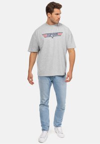 Šedé bavlněné tričko s grafickým nápisem "TOP GUN" v modré a červené barvě, kombinované se světle modrými džíny a bílými teniskami. Krátké rukávy a uvolněný střih.