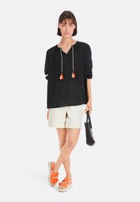 Schwarze leichte Bluse mit Quasten, kombiniert mit cremefarbenen Shorts, orangefarbenen Sandalen und einer schwarzen geflochtenen Handtasche.
