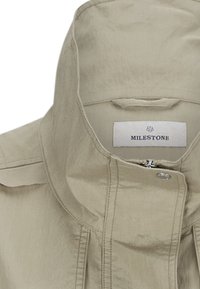 Helle beige Jacke mit hohem Kragen, Reißverschluss und Druckknopf. Glattes Material mit sichtbaren Nähten und einem Etikett mit der Aufschrift "MILESTONE."