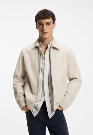 Joven con cabello rizado que lleva una chaqueta beige con cremallera sobre una camisa ligera a rayas y pantalones oscuros, de pie con las manos en los bolsillos.