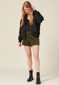 Schwarze Bomberjacke mit gerippten Bündchen, kombiniert mit olivgrünen Cordshorts. Modell trägt schwarze klobige Knöchelstiefel. Hintergrund beige.