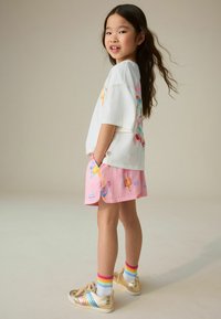 Jong meisje met lang donker haar dat een wit shirt met print draagt, roze korte broek met kleurrijke prints, regenboogsokken en gouden sneakers, staand zijwaarts.