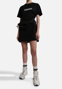 Person med en sort Napapijri grafisk t-shirt, sort nederdel, sort bæltetaske, hvide sokker med striber og hvide højhælede sneakers.