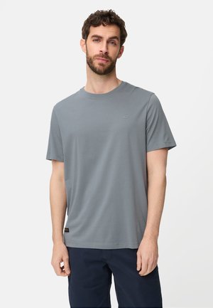 Brodaty mężczyzna ubrany w zwykły szary T-shirt z krótkim rękawem i ciemne spodnie, stojący na białym tle.