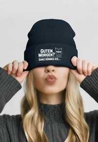 Black knit beanie with a wide band featuring white text and an owl graphic. Model has long, wavy hair and wears a gray ribbed sweater.