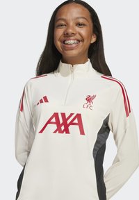 Leende ung person med långt hår som bär en vit Liverpool FC träningströja med röda logotyper och svarta sidopaneler.