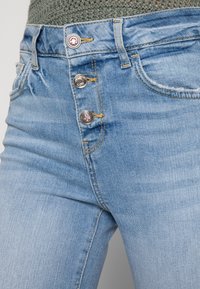 Jeans de ganga azul claro com um design de cintura alta, fecho de três botões e detalhes de costura subtis perto dos bolsos.
