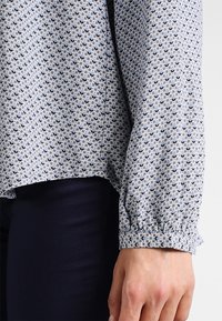 Vaaleansininen kuvioitu paitapusero, jossa on pieniä tummansinisiä kukkamotiiveja. Ominaisuuksiin kuuluu joustava hihansuu ja hieman rento istuvuus, yhdistettynä tummiin housuihin.