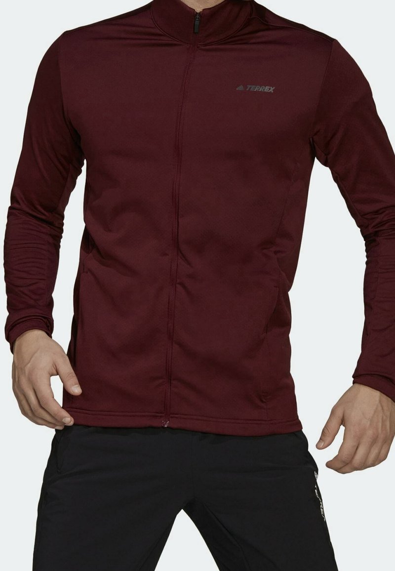 Chaqueta de cremallera de manga larga burdeos en un tejido suave y texturizado, con puños ajustados y un logo negro en el pecho. Diseño minimalista, corte atlético.