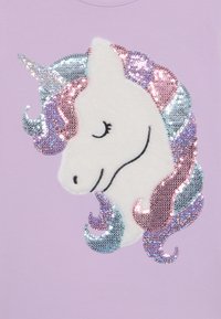 T-shirt lilla con design di unicorno, caratterizzato da un volto bianco peloso e una criniera arcobaleno in paillettes con tonalità metalliche di rosa, blu e viola.