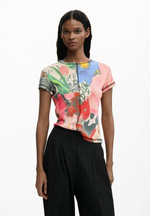 Femme aux longs cheveux foncés portant un haut à manches courtes avec motif floral et un pantalon noir taille haute à jambes larges, debout devant un fond blanc.