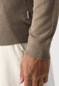 Brązowy robiony na drutach sweter z prążkowanymi mankietami, z delikatną teksturą; metka z boku pokazuje nazwę marki. Ręka znajduje się w pobliżu.