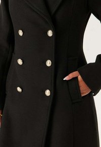 Cappotto di lana nera con design doppiopetto, bottoni argentati, colletto a revers e tasche laterali. Textura liscia con vestibilità su misura.