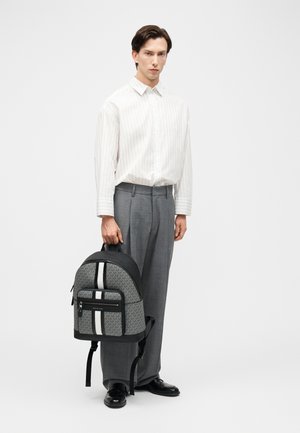 Bărbat purtând o cămașă albă cu dungi și pantaloni gri, ținând un rucsac gri cu model dungi albe și negre.