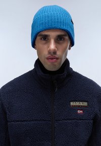 Blå stickad mössa med ribbat mönster sitter på en persons huvud. De bär en mörk marinblå fleecejacka med en logomärke på bröstet.