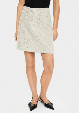 Falda mini de color beige claro con una superficie texturizada, que presenta acentos metálicos y un diseño clásico de cinco bolsillos. Combinada con bailarinas negras de punta.