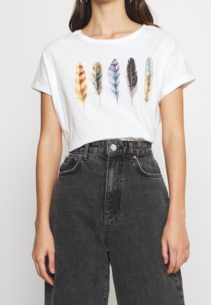 Kobieta w białym t-shircie z pięcioma kolorowymi piórami nadrukowanymi na piersi i czarnych, wysokich jeansach.