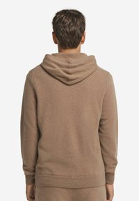 Mann in einem hellbraunen Kapuzenshirt und passenden Hosen, mit dem Rücken zu einem schlichten weißen Hintergrund gewandt.