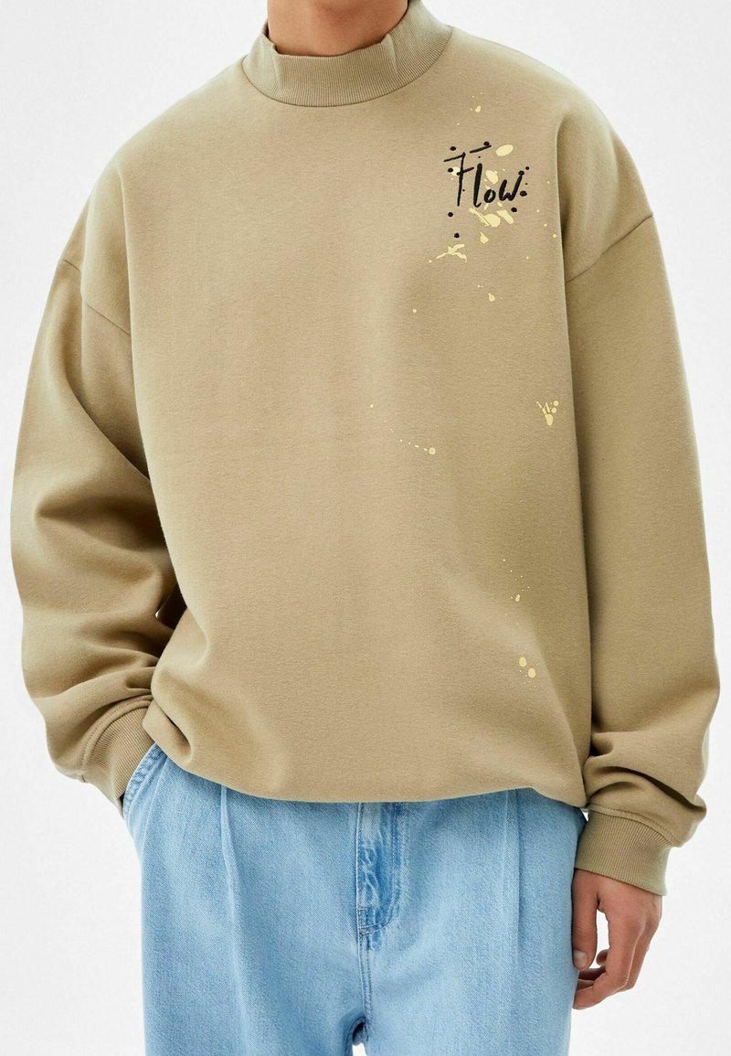 Kaki sweatshirt med hög krage, med en svart "Flow." tryck och gula färgstänk detaljer. Bärs med ljusblå denimbyxor.