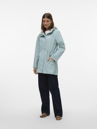 Nainen pukeutuneena vaaleansiniseen hupulliseen sadeviittaan, kädet taskuissa, tummat farkut ja ruskeat kengät, seisomassa valkoista taustaa vasten.