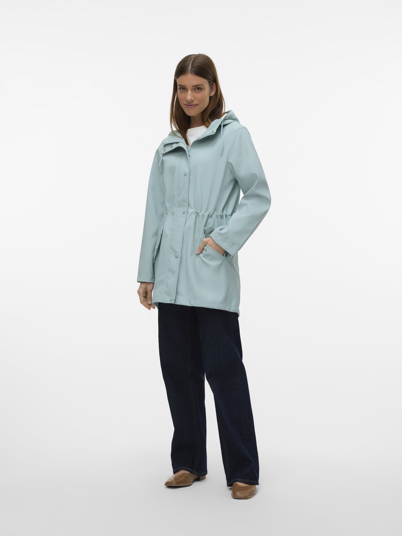 Nainen pukeutuneena vaaleansiniseen hupulliseen sadeviittaan, kädet taskuissa, tummat farkut ja ruskeat kengät, seisomassa valkoista taustaa vasten.