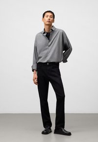 Camicia grigia con bottoni e vestibilità rilassata, abbinata a jeans neri a gamba dritta. Scarpe slip-on nere completano il look. Texture del tessuto lisce.