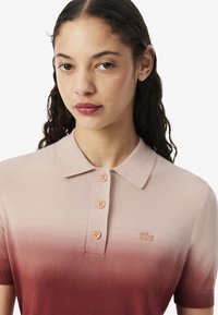 Polo shirt dégradé présentant une transition de couleur du rose clair au bordeaux, à manches courtes, avec un col et quatre boutons pêche ornés d'un logo sur la poitrine.