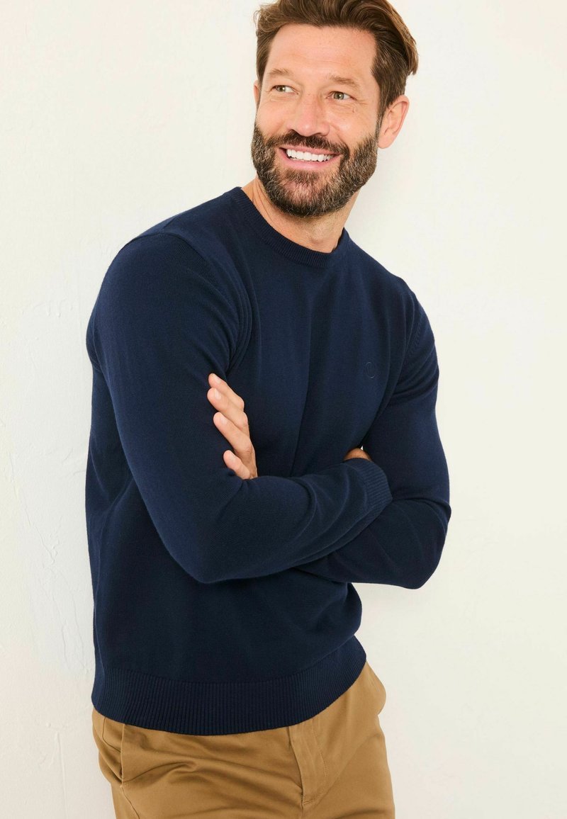 Marineblaue Crewneck-Pullover aus weichem, strukturiertem Strick mit gerippten Bündchen und Saum, kombiniert mit hellbeigen Hosen. Keine sichtbaren Logos.