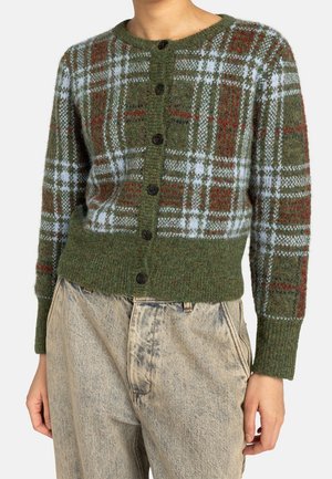 Zelený kardigan na knoflíky s červeno-bílým kostkovaným vzorem, nošený s vysoce pasovanými světlými béžovými džínami s efektem acid wash.