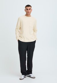 Camicia a maniche lunghe color crema con taschino frontale, abbinata a pantaloni neri e scarpe bianche con strisce nere. Fit casual e rilassato.