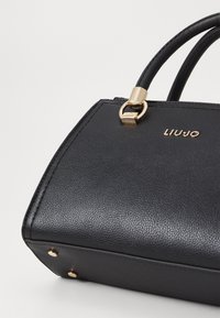 Bolso de mano de cuero negro con acabado texturizado, herrajes dorados, asas redondeadas y nombre de marca en relieve. Presenta tachuelas doradas en la base.