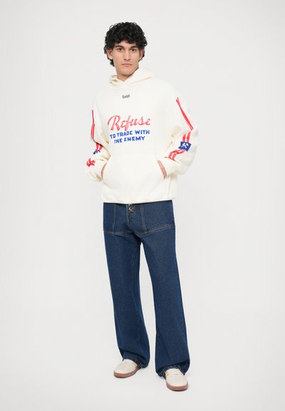 Light cream hooded sweatshirt with blue and red accents and text; paired with loose-fitting dark blue denim jeans and white sneakers.