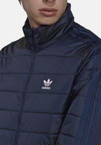 Giacca puffer blu navy con zip, con il logo Adidas sul petto, motivo a righe orizzontali e tre strisce sulle spalle.