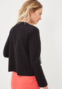 Chaqueta negra recortada con mangas largas, escote redondeado y aberturas laterales. Textura de tela suave, combinada con una falda coral.