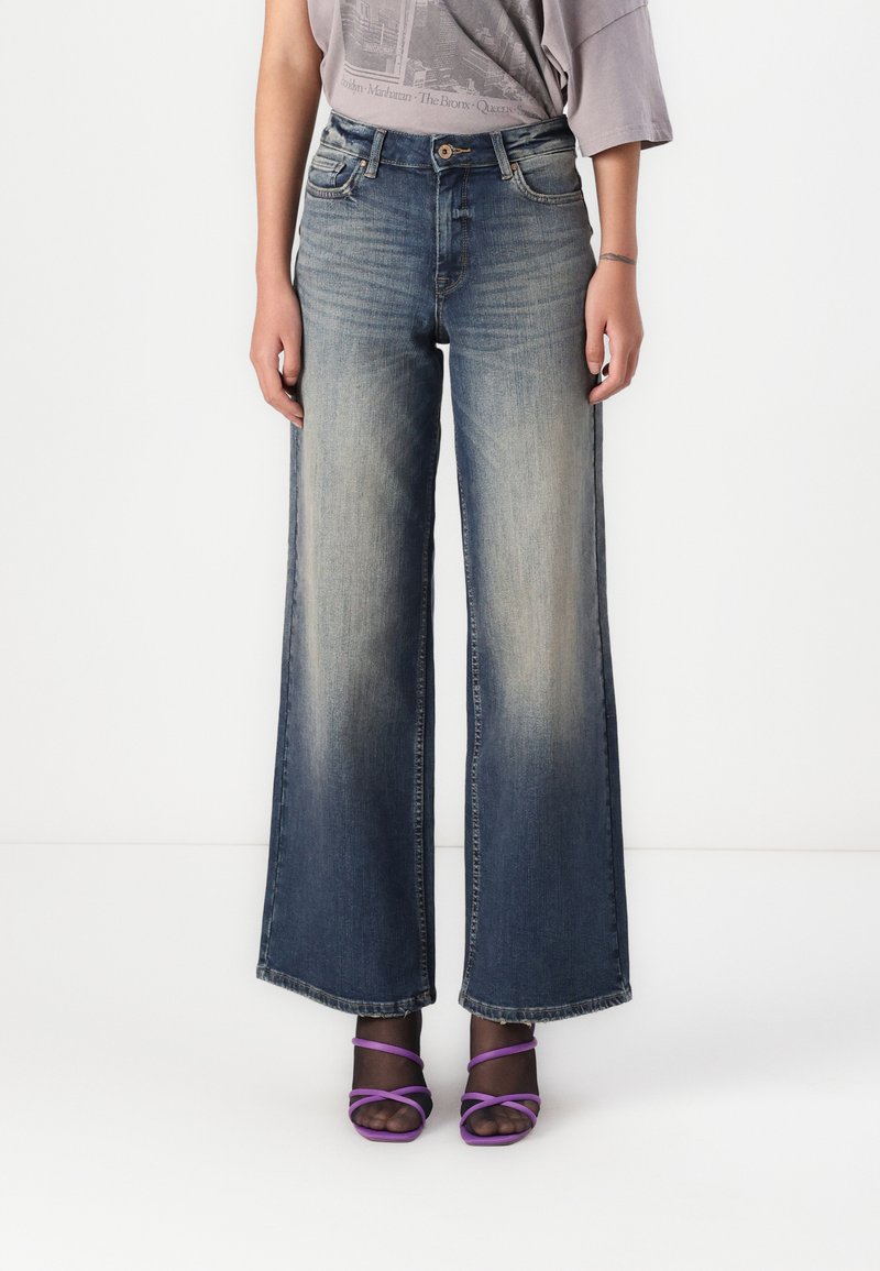 Bredde ben, falmet blå denimbukser med høyt liv, fem-lomme design og kontraststing. Brukt sammen med lilla sandaletter med høye hæler.