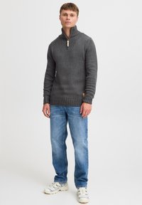 Grå strikket sweater med høj krave, prydet med ribbede detaljer og en træzipper. Sammen med lyseblå jeans og hvide sneakers.