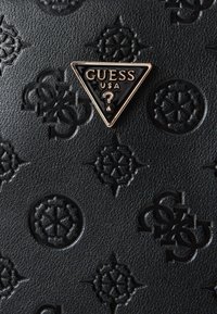 Musta tekstuuripinnaga materjal, millel on reljeefsed mustrid. Seal on kolmnurkne kuldtooniga logo, millel on kiri "GUESS USA" ja küsimärgi sümbol.