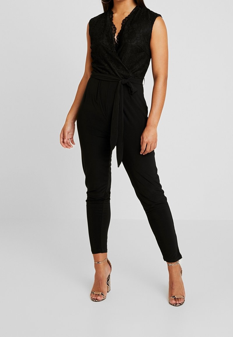 Femme portant une combinaison noire sans manches en dentelle à nouer à la taille avec pantalon noir ajusté, associée à des sandales à talons hauts en cuir façon peau de serpent.
